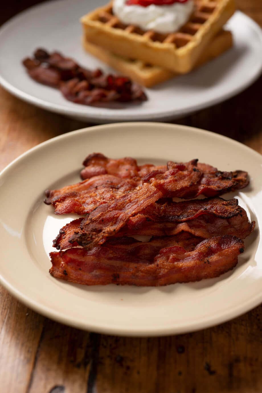 Bacon on a white plate with waffles in the background